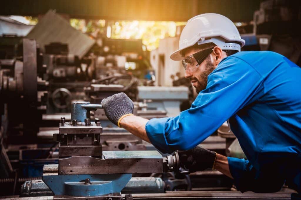 Ein Facharbeiter in blauer Arbeitskleidung und Schutzhelm bedient eine CNC-Maschine in einer industriellen Werkhalle. Der Arbeiter nutzt präzise Einstellungen an der Maschine und trägt dabei Sicherheitsbrille und Handschuhe. Im Hintergrund sind weitere Maschinen und Werkzeuge sichtbar.