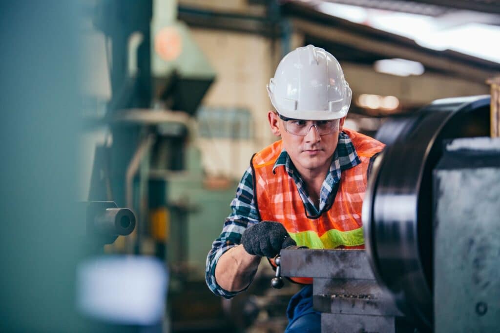 Ein konzentrierter Arbeiter mit Schutzhelm und Sicherheitsweste bedient eine CNC-Maschine in einer industriellen Umgebung. Er justiert präzise die Maschine, während er durch Schutzbrille und Handschuhe zusätzlich abgesichert ist. Im Hintergrund sind weitere Maschinen und Produktionsanlagen zu erkennen.